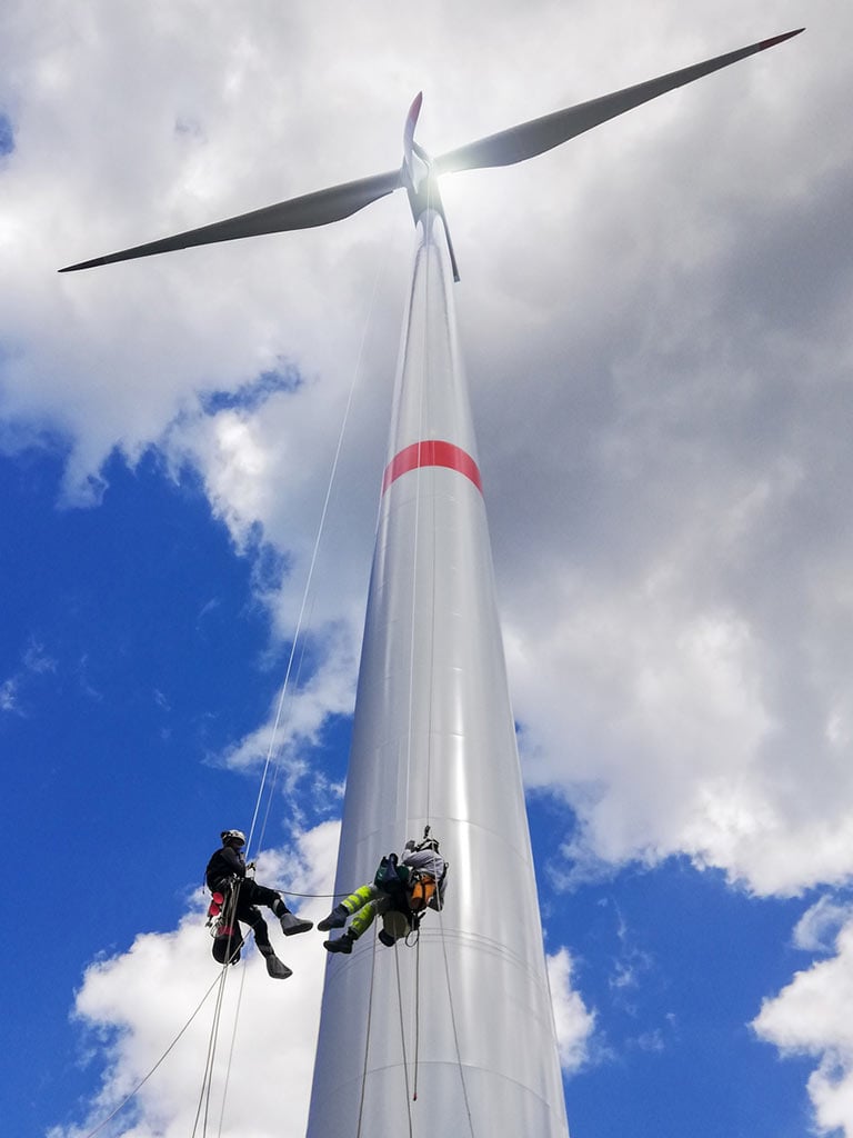 View from bottom on wind-turbine and two rope access technicians rappelling down from blade on the ropes